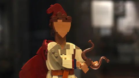Corinium Museum A Lego figure of Orpheus, who is wearing a red cloak and hat and is holding a lyre. He has brown hair.