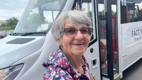 John Devine/BBC Janet Wallis has short grey hair and dark framed glasses on she is wearing a colourful raincoat with a hood that is down, behind her is a white mini bus.