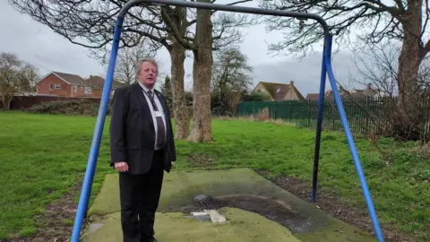 North East Lincolnshire Council A man in a black suit and tie with a white shirt and a lanyard is took next to a damaged play area apparatus. The green tarmac belong the metal bars of a swing has been scorched black.