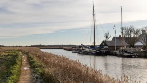 Suffolk County Council A view of a river and field running alongside it. Some boats have been moored on the opposite side of the river near some buildings. 
