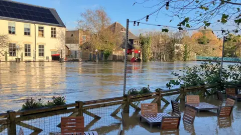 Chairs and tables are completely submerged in murky flood water after the River Avon burst its banks. You can see flood water behind which stretches across the town.