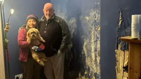 Phil Boulton Phil and Michelle Boulton, who is holding their pet dog Amber, are pictured in a room with large white marks on the wall. Phil is wearing a dark fleece and white trousers, while Michelle is wearing a pink hat and a pink jacket.