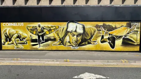 Nathan Murdoch Street Arts Hire Peterborough Mural depicting four images of Walter Cornelius in yellow, black and white. On the right, he is seen on his hands and knees in front of a canon ball; next he is running along a river bank with a bridge behind him and wearing a pair of striped wings; next he is face on, flat on his stomach, with his hands beneath his shoulders, looking up and in front of him is a pea; and the final image shows him falling face down into a river while wearing wings. 