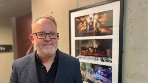 Jimmy Fay wearing a gray blazer and black shirt, he is wearing black glasses and has white hair