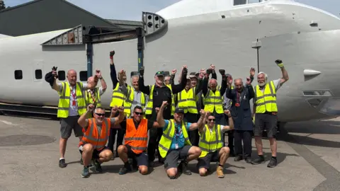 A group of people wearing high vis jackets holding their arms up and cheering, photographed in front of a large plane