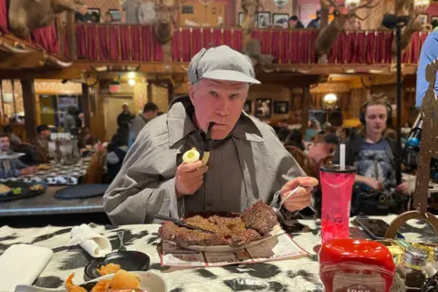 Netflix Will Ferrell wearing a Sherlock Holmes costume - a dogtooth cape, matching hat and a pipe - in front of a huge steak on a plate in a restaurant