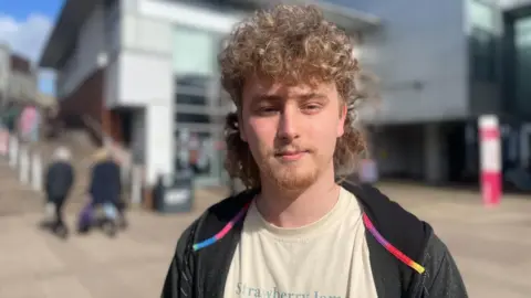 Simon Thake A man with a curly blonde mullet haircut stands squinting slightly in the sunshine