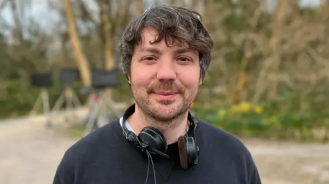 Simon Mayhew-Archer on set of Can You Keep a Secret? He has greying hair and is wearing a black jumper.