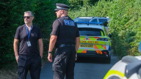 PA Media A police officer and another person stand on a narrow country lane beside a marked police car. Dense green hedges line both sides of the road and additional police vehicles are parked further along the lane.