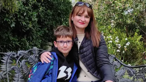 Family photo A portrait photo of Dima and Elena staring at the camera while sitting on a park bench. He is waring a blue jacket and has glasses. She is wearing a black jacket and white jumper and has sunglasses on her head.