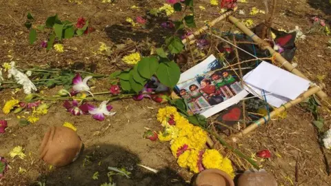 Yellow flowers, and pink and white flowers and pots are lying on soil next to a photo of a young woman standing with children. 