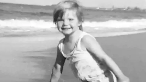 Grimmer family A black and white photo of a blonde girl in a swimsuit on a beach