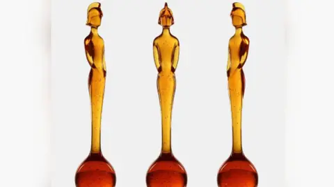 BRIT Awards Três estátuas idênticas são exibidas lado a lado contra um fundo branco. Cada estátua é feita de material translúcido de cor âmbar com pequenas manchas escuras por toda parte. As estátuas têm forma alongada e elegante, lembrando uma figura humana estilizada em pé com os braços próximos ao corpo. A base de cada estátua tem uma forma arredondada em forma de bulbo que gradualmente se estreita em um torso e cabeça altos e esguios. As figuras possuem um capacete em forma de capacete, o que lhes confere uma silhueta distinta.