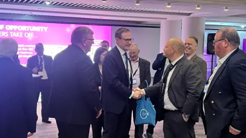 BBC A man with glasses and a suit smiles as he shakes hands with another man in a suit at the tory conference. They are surrounded by other men attending the conference. 