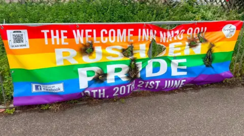 A colourful Pride banner, in front of a bush, that has about eight burnt holes in it. There is a path in front. It is promoting a Pride event. 