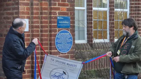 Two men pulling a red white and blue banner to reveal a blue plaque on a red brick wall.