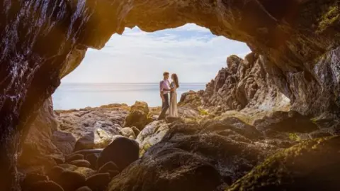 Ben Fiore A photo showing a bride and groom facing eachother with the sea behind them, viewed from a rocky cave aperture.