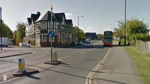 A residential road junction with a large building on the far side and a red bus in the distance