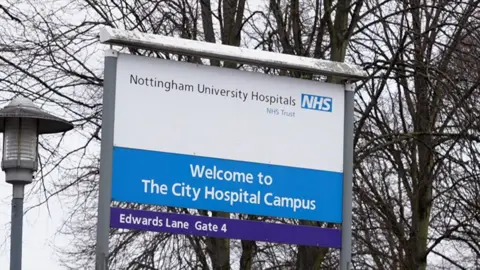 Tanda biru dan putih di luar Rumah Sakit Kota Nottingham yang mengatakan: Selamat datang di kampus Rumah Sakit Kota, Nottingham University Hospitals NHS Trust. Ada lampu jalan di sebelah kiri tanda dan pohon tanpa daun, di belakang tanda.