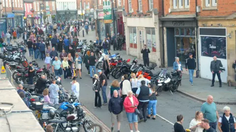 South Kesteven District Council Grantham Bike Night in 2025. The picture shows dozens of motorbikes parked at both sides of a road. Dozens of people are walking down the road looking at the bikes.