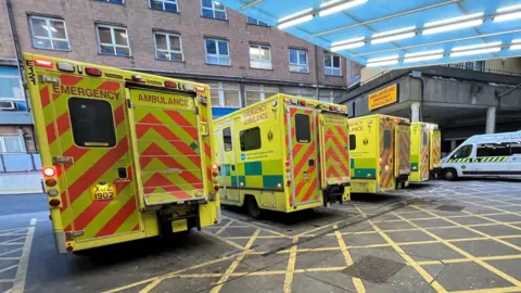 The outside of an Emergency Department main entrance. 5 ambulances are parked outside.