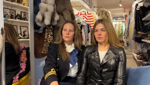 Two people are sitting side by side inside a vintage clothing shop. They are surrounded by racks of colourful garments, including fur coats, and accessories are displayed on shelves and hangers.