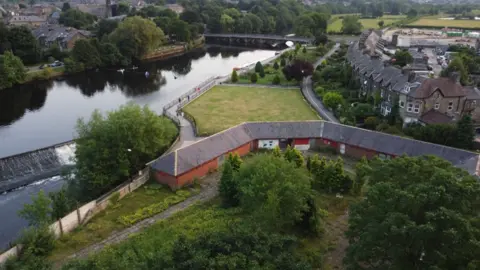 Aerial shot of the lido. The clubhouse and changing rooms are still standing yet the area where the pool sat, is now covered in grass and trees. The neighbouring River Wharfe is seen to the left hand side.