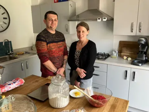 BBC A man wearing a jumper is on the left next to a woman with dark hair wearing a dark top. They are in the kitchen looking at the camera. Cupboards are behind them and a table, with items on, is in front of them.