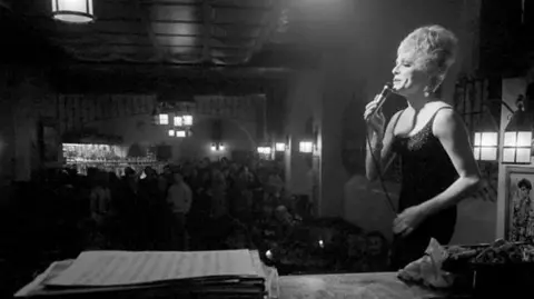Watford/Mirrorpix/Getty A black and white photo of drag queen Shane performing downstairs at The Black Cap. She is stood on the stage on the right, wearing a strapless dress, hair up in a chignon, and in the background is the audience. She is holding a wired mike and the spotlight is shining on her.