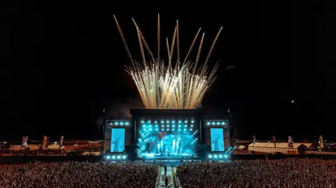 Boardmasters Festival / WillBaileyPhoto hundreds of people stand in a crowd watching a performer on stage at night time, with fireworks bursting out into the sky from behind the stage.