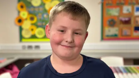 A young boy looks into the camera. He has fair hair and is wearing a dark top. There are brightly coloured pictures on the wall behind him.