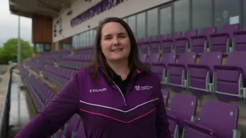 A woman stood in the middle of football stadium seats in a purple Loughborough University jumper, smiling at the camera.