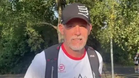 Nigel Watson A man in a white t-shirt and black cap, with black rucksack straps over his shoulders. He is wearing a serious expression and looking at the camera. He has a white beard and is standing outside, with green trees behind him.