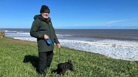 Amanda White / BBC A man standing on top of grass with the sea in the background. He is walking a small black dog on a lead and is wearing black trousers, a green rain jacket and a black bobble hat.