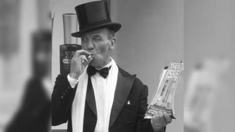 B&w photo of Mr Walker in top hat and tails holding the Big Issue magazines and smoking a cigarette