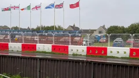 MANX RADIO A brown wooden fence and white and red alternating crash barriers run parallel either side of the start-finish straight of a very wet Billown Course. A row of flag poles featuring the Manx, British, Scottish, English, Irish, and Welsh flags flying stiffly in the wind sits behind the barriers. There is also fencing in place in front of cars parked on a grassed area.