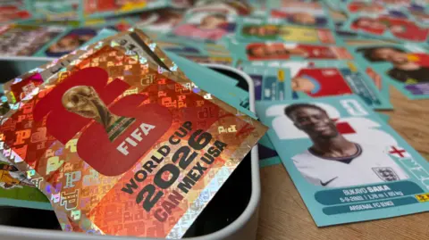 Panini stickers scattered on a table with two stickers in the foreground, one showing a picture of the World Cup trophy, the other showing England winger, Bukayo Saka wearing the white England home shirt.