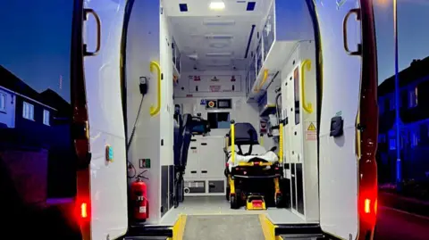 The back of an ambulance with its doors open, showing a bed for a patient inside and cupboards around the edge of the interior. There are houses in the background.