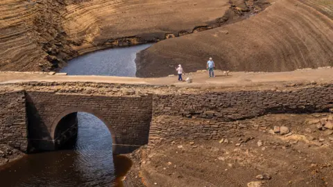 PA Media Two walkers go over a bridge usually submerged by water in Yorkshire. A dry, clay like landscape is around them with a small trickle of water going under the bridge they're walking on.