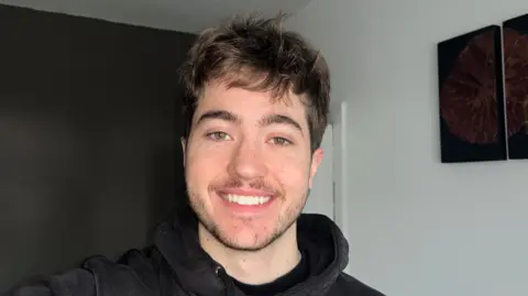 Ethan Dauncey Ethan Dauncey is wearing a black hoodie and smiling. He has brown hair and facial hair. Behind him is a black wall and a white wall.