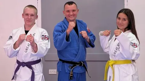 Draco Dojo A teenage boy and a teenage girl are pictured in jiu-jitsu clothing with green and purple belts. They are standing either side of their sensei, who is wearing a dark blue uniform.