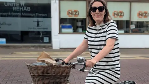A woman is wearing a white and navy striped dress and shades. She is sitting on a bike with a brown basket on the front. 