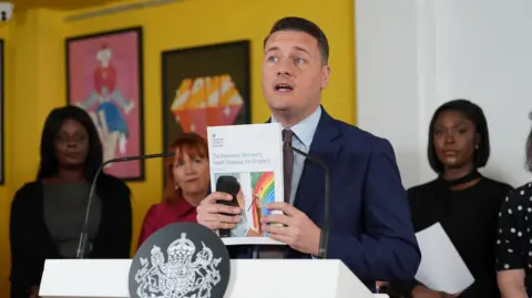 PA Media Wes Streeting holds up the published strategy report while speaking at a lectern branded with the government emblem. A number of women stand in the background listening to him.