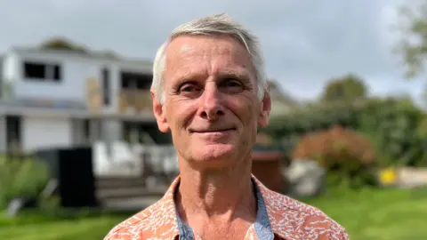 A man with grey hair smirks at the camera, wearing a patterned orange and white top.