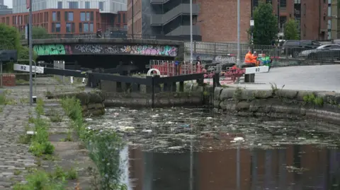 The canal showing low levels of water. It has rubbish floating on the surface of the water.