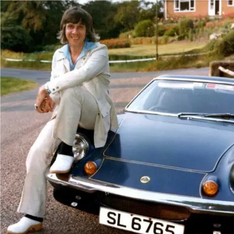 Stan Laundon Stan Laudan, who in the picture has shoulder length brown hair, a blue shirt, white suit and white shoes and is sitting on a dark Lotus car in the 1970s.