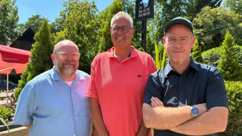 Three men in blue, pink and navy shirts smile at camera in front of Black Horse sign
