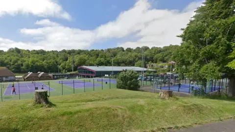 A sports complex with racquet sports courts and a pavilion. The complex is surrounded by grass and trees.