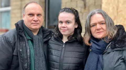 Nicola Rees/ BBC A middle-aged man and woman - both with grey hair - stand in front of a boarded-up house, alongside a young girl who has long, dark hair. They all wear dark-coloured coats and scarves, and there is rain on their jackets.