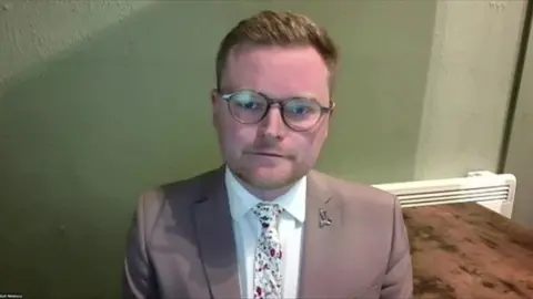 BBC A man with light brown hair and brown glasses is sitting on a chair in front of a green wall. He is wearing a white shirt, brown suit jacket and a white, pink and green floral tie. There is a silver pin on his lapel
